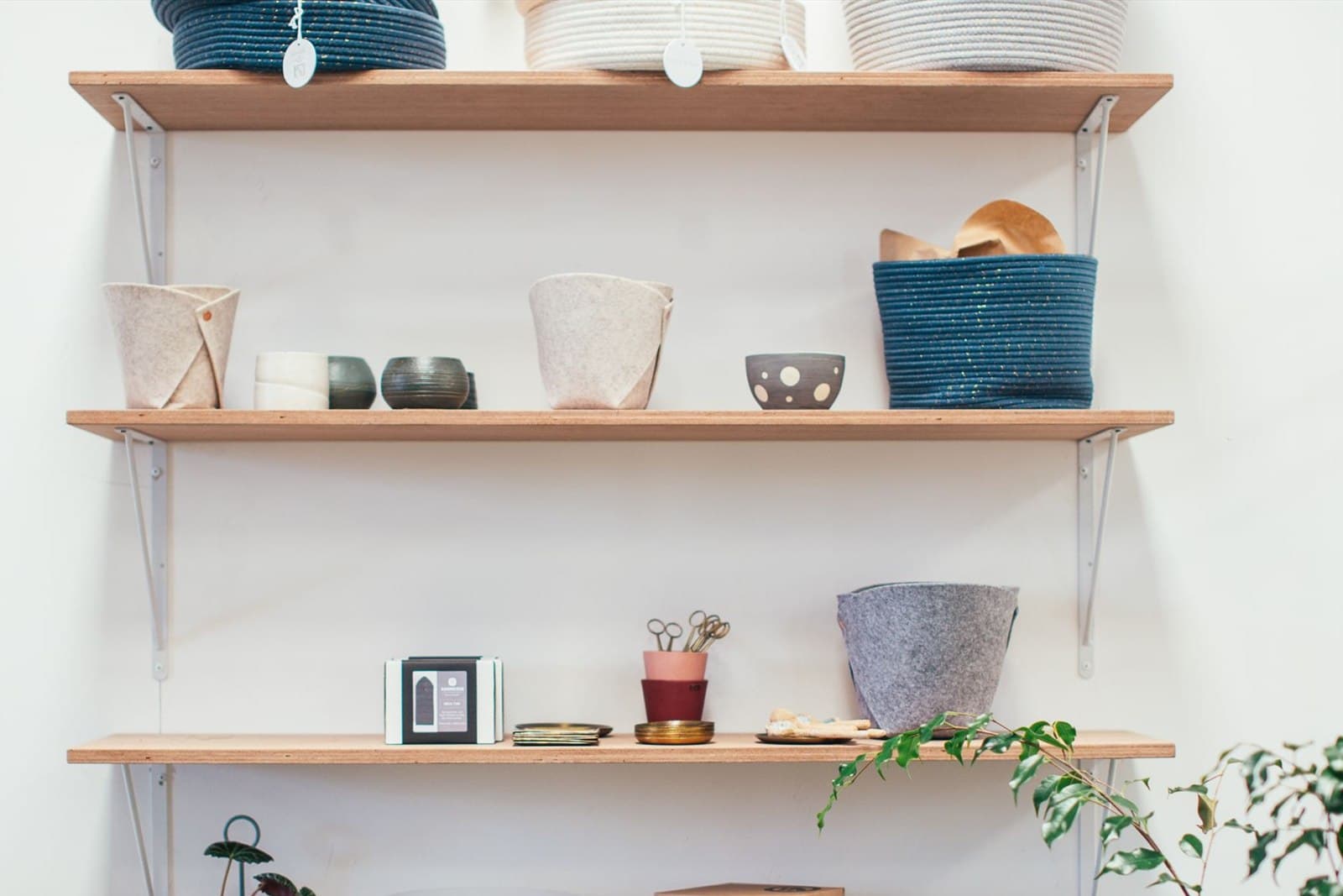 Three plywood shelves hold felt pots, ceramic bowls and woven baskets against a white wall