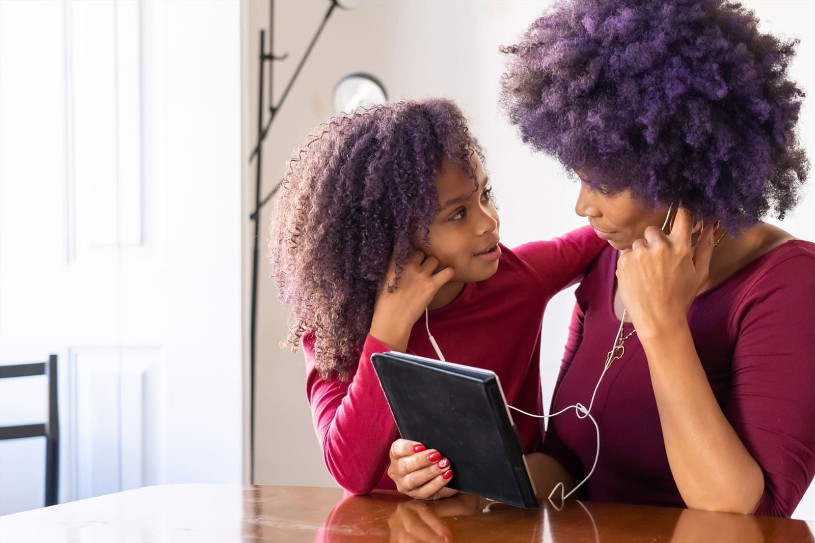 A girl and a woman share a pair of earphones at a table, watching a tablet