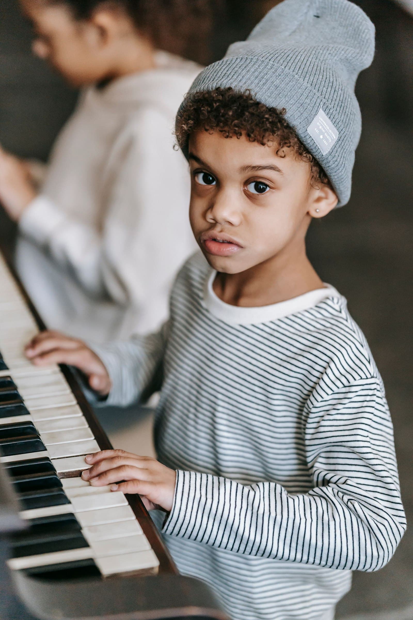 A boy in a grey beanie rests his hands on the keys of an upright piano