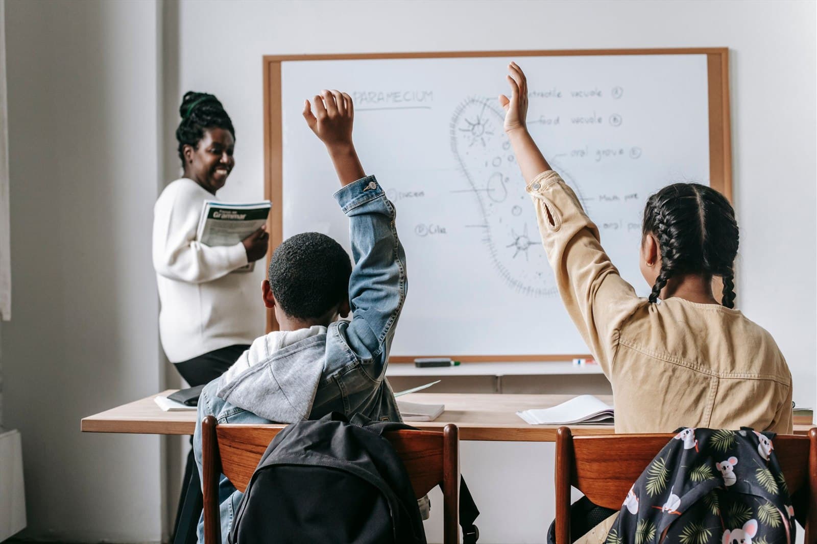 Two pupils raise their hands at a shared desk while their teacher stands by the whiteboard
